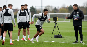 Professional football players using wearable tracking vests and data tablets on the pitch.